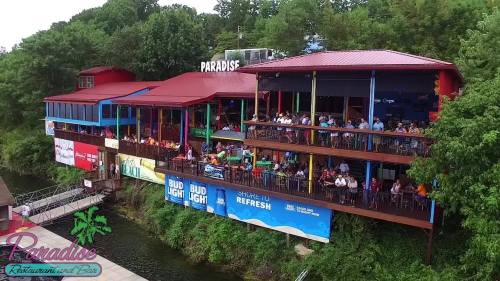 A vibrant restaurant by the water, featuring outdoor seating and a lively crowd, surrounded by greenery.
