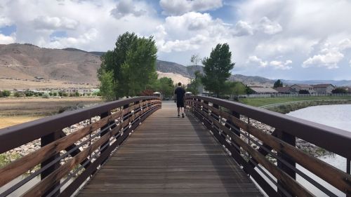 A person walks on a wooden bridge surrounded by trees and mountains under a partly cloudy sky.