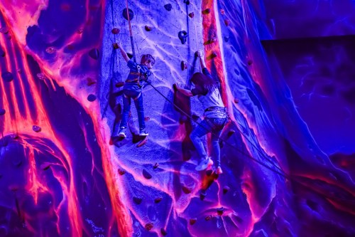 Two children climbing a colorful rock wall with vibrant, abstract patterns in the background.