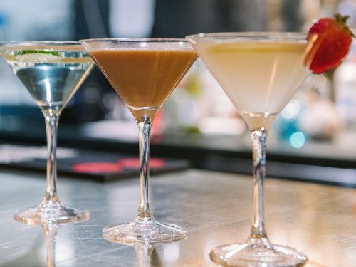 Three elegant cocktails in martini glasses: a blue drink with lime, a brown drink, and a yellow drink with a strawberry garnish.