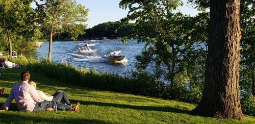 People relax on a grassy shore while boats speed across a calm lake under a clear blue sky.