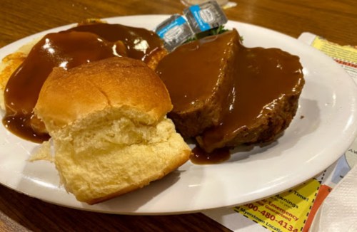 A plate with meatloaf covered in gravy, a roll, and two packets of butter on a wooden table.