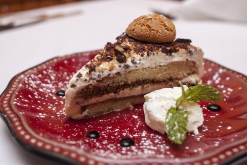 A slice of layered cake topped with cream, chocolate shavings, and a cookie, served on a decorative red plate.