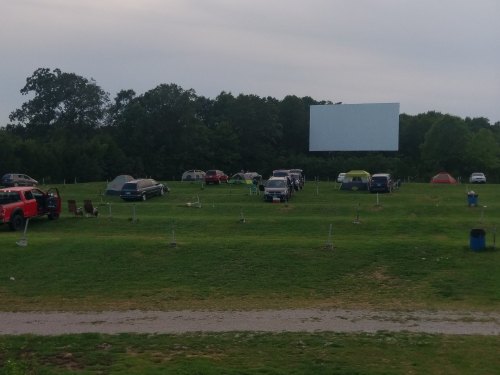 A grassy area with parked cars and tents, featuring a large blank screen in the background, likely for a drive-in movie.