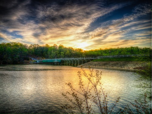 A serene sunset over a river, with a bridge in the foreground and lush trees lining the banks.