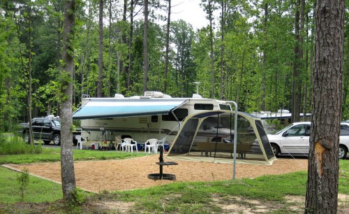 A camper parked in a wooded area, with a tent and picnic setup nearby, surrounded by trees and parked cars.