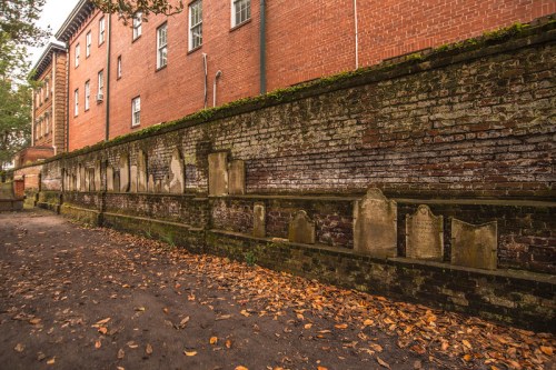 A weathered brick wall lined with old gravestones, surrounded by fallen leaves and historic buildings.