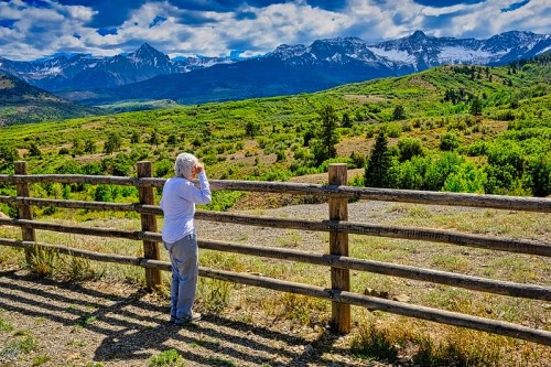 A person stands by a wooden fence, gazing at a scenic mountain landscape under a bright blue sky.