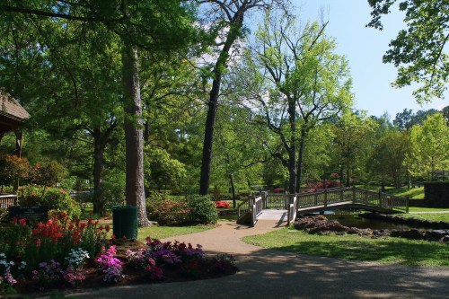 A serene park scene featuring a wooden bridge over a pond, surrounded by lush greenery and colorful flowers.