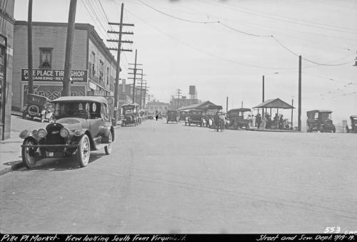 Black and white photo of a street scene with vintage cars, shops, and people walking along the waterfront.