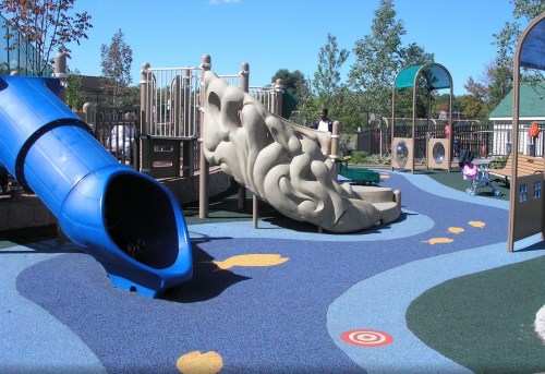 A colorful playground featuring a blue slide, climbing structure, and soft rubber flooring under a clear blue sky.