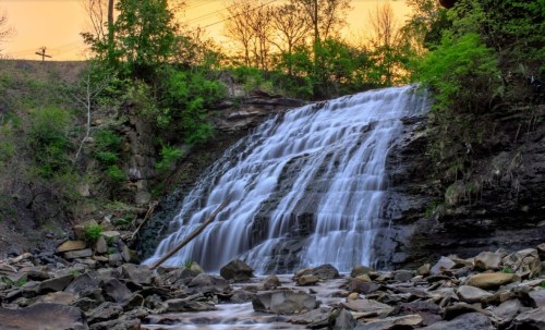 A serene waterfall cascades over rocks, surrounded by lush greenery under a warm sunset sky.