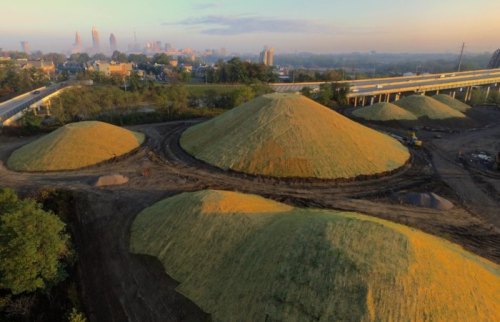 Aerial view of large, grassy mounds in an industrial area, with a city skyline in the background and morning light.