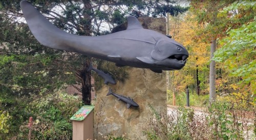 A large fish sculpture mounted on a stone, with smaller fish below, surrounded by trees in autumn colors.