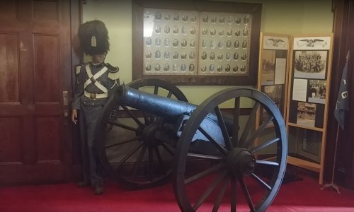 A historical display featuring a soldier in uniform next to a large cannon, with framed photos on the wall behind.