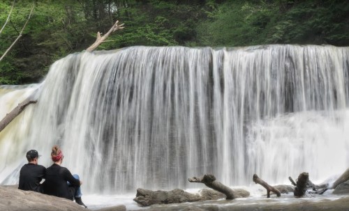 A couple sits on a log by a waterfall, surrounded by lush greenery and flowing water.