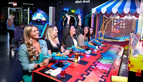 A group of five women joyfully playing arcade games, holding toy guns, with colorful lights and attractions in the background.