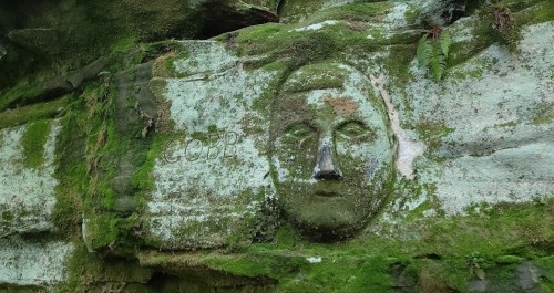 A moss-covered rock face with a carved human-like face, surrounded by greenery.