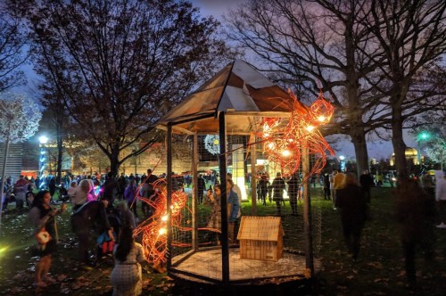 A park scene at dusk with a lit structure, surrounded by people enjoying an outdoor event among trees.