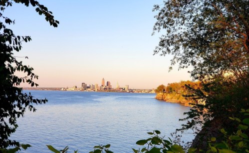 View of a city skyline across a calm lake, framed by trees and bathed in warm sunset light.