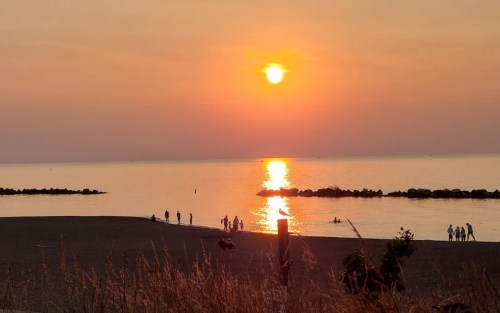 Sunset over a calm beach, with silhouettes of people walking along the shore and reflecting light on the water.