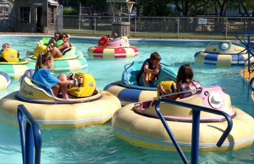 Children enjoying bumper boats in a pool on a sunny day, surrounded by colorful inflatable boats.