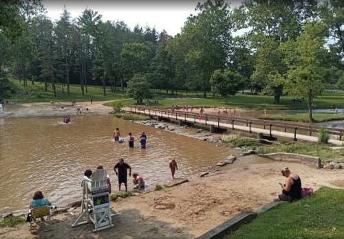A sandy beach area by a calm lake, with people swimming, lounging, and enjoying the outdoors on a sunny day.