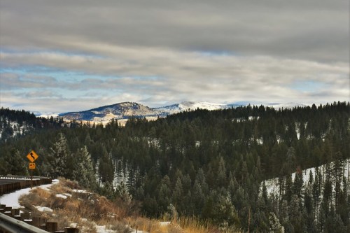 A winding road through a snowy landscape, surrounded by evergreen trees and distant mountains under a cloudy sky.