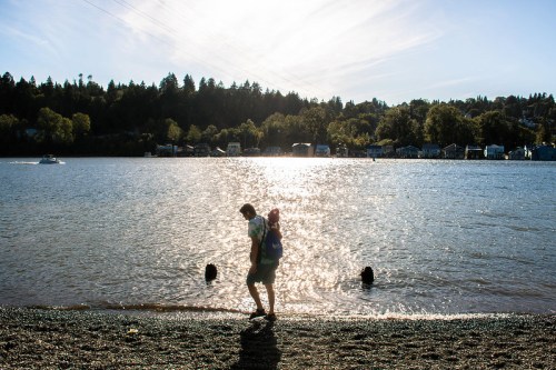 A silhouette of two people walking along a beach by a river, with trees and houses in the background under a bright sky.