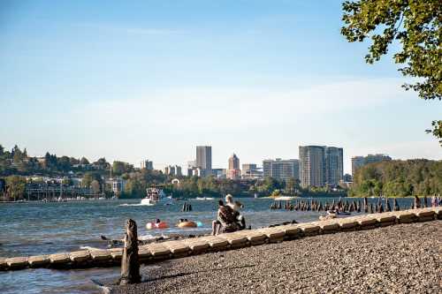 A couple sits on a beach by the water, with a city skyline and boats in the background under a clear blue sky.