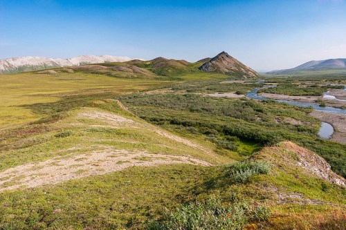 A scenic landscape featuring rolling green hills, a winding river, and distant mountains under a clear blue sky.
