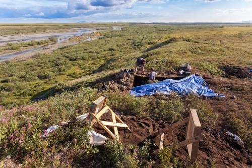 Two people excavate a site near a river, surrounded by lush greenery and a blue sky with scattered clouds.