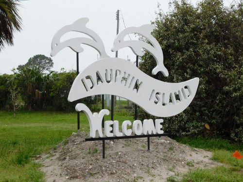 Welcome sign for Dauphin Island featuring two dolphin silhouettes above the text. Greenery in the background.