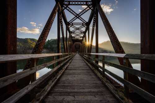 A wooden bridge stretches over calm water, framed by mountains and a sunset in the background.