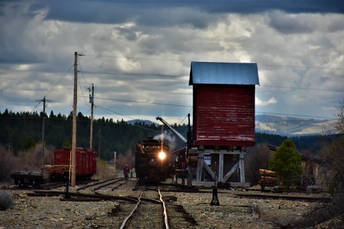 A vintage steam train approaches a red wooden water tower on a cloudy day, with rail cars and trees in the background.
