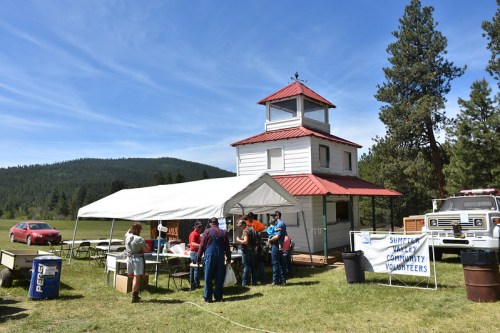 A community event with a white building and a tent, surrounded by trees and mountains, with people gathered outside.