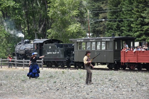 A woman in a blue dress gestures dramatically while a man with a rifle stands nearby, with a steam train and passengers in the background.
