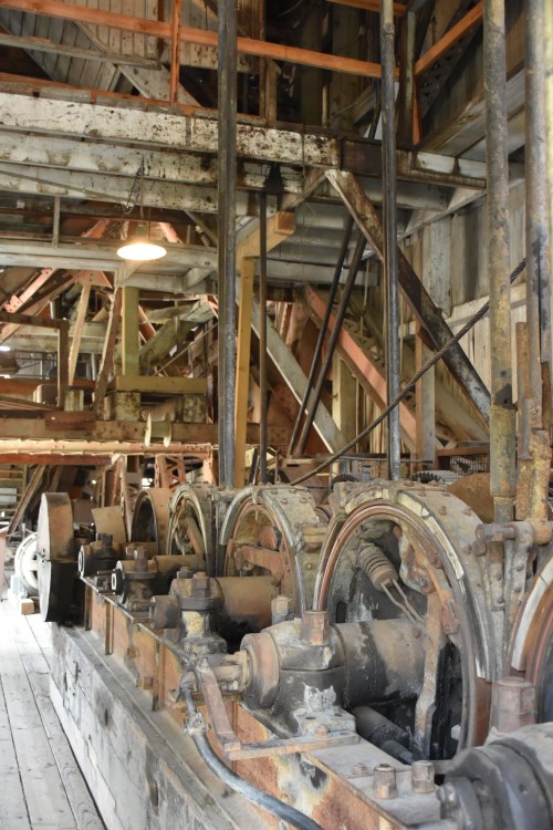 Interior of an old industrial building featuring large, rusted machinery and gears, with wooden flooring and beams.
