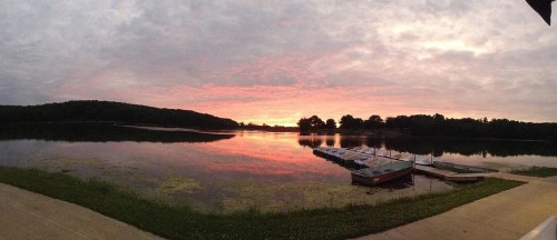 A serene lake at sunset, reflecting vibrant colors in the water, with boats docked along the shore and trees in the background.