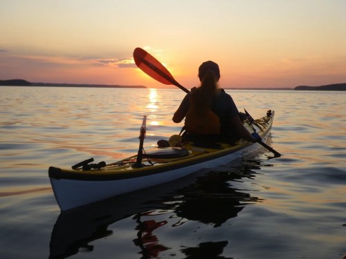 A person kayaking on calm water at sunset, with vibrant colors reflecting on the surface.