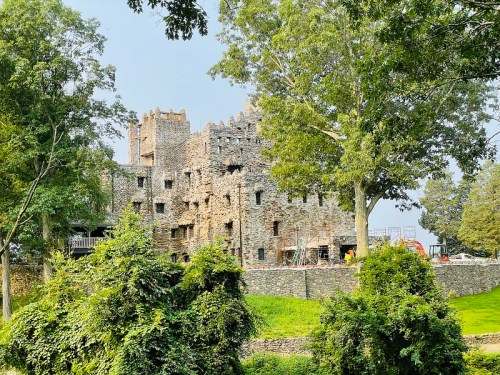 Ruins of a stone castle surrounded by trees and greenery, with construction equipment visible in the background.