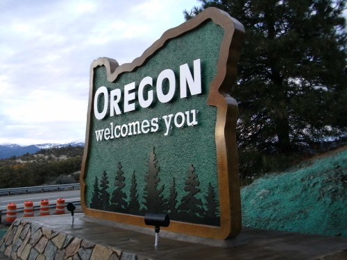 Sign welcoming visitors to Oregon, featuring the state outline and trees, with a cloudy sky in the background.