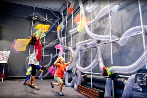 Children play with colorful fabric pieces in a hands-on exhibit featuring tubes and interactive elements.