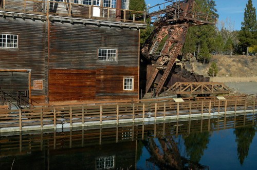 A weathered wooden building beside a calm waterway, featuring a large, rusted machinery structure.