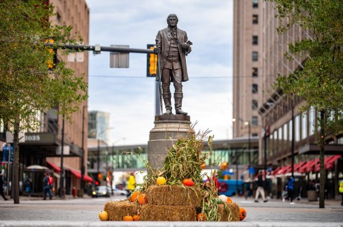 A statue of a historical figure stands on a street, surrounded by pumpkins, hay, and autumn decorations.