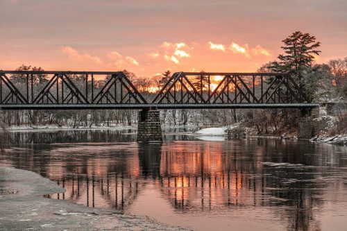 A metal bridge spans a river at sunset, reflecting vibrant colors on the water's surface, surrounded by trees.