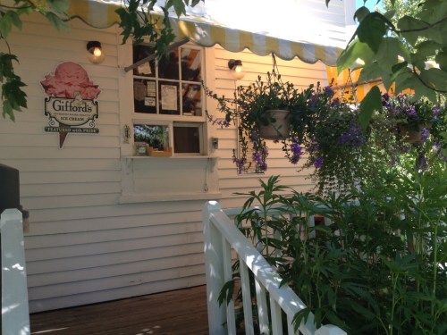 A charming ice cream shop with a white exterior, yellow awning, and flower pots, surrounded by greenery.
