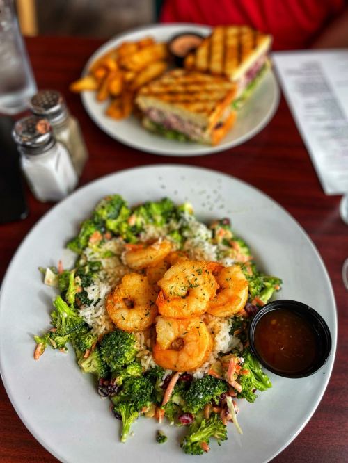A plate of shrimp over rice and broccoli, with a side of dipping sauce, alongside a sandwich and fries in the background.