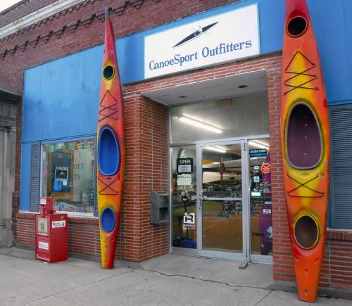 Colorful kayaks flank the entrance of CanoeSport Outfitters, a shop with a welcoming "Open" sign.