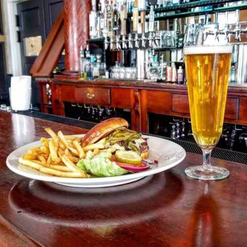 A plate of fries and a burger with lettuce and tomato, alongside a tall glass of beer, set on a bar counter.